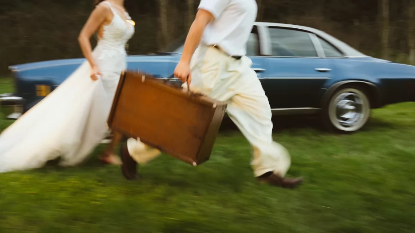 Çimenlikte elinde eski tip deri bavul ile koşan gelin ve damat; arkada mavi klasik otomobil
